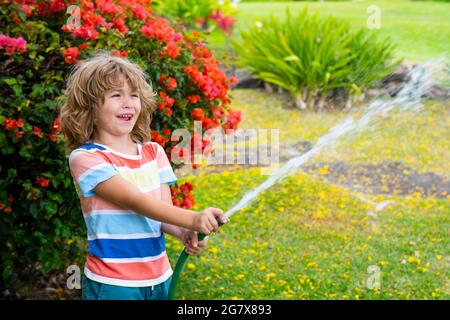 Cute little boy watering flowers in the garden at summer day. Child using garden hose. Funny kid watering plants in the yard garden. Stock Photo