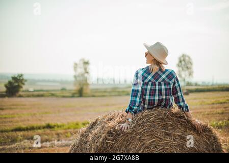 Woman in hat sitting on round bales in the field, enjoying good weather. Beautiful landscape for young entrepreneurs farmers. Back view image in the s Stock Photo