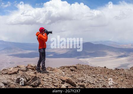 A young woman on the top of mountain in action of photographing the beauty of volcanic landscape in Parque Nacional Nevado de Tres Cruces near Copiapo Stock Photo