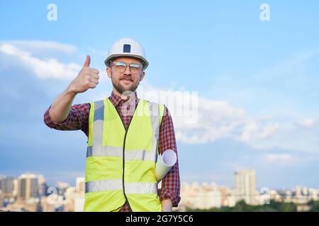 Attractive bearded male engineer or construction architect in helmet and safety reflective vest showing thumbs up gesture. Man holding blueprints in sunny day blue sky and urban city skyline Stock Photo