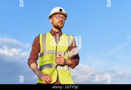 Bearded male supervisor or construction architect in helmet and safety reflective vest with tape-measure on a construction site. Man holding blueprints in sunny day blue sky and urban city skyline Stock Photo