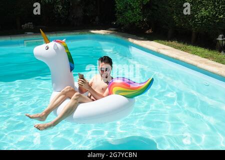 Young man wearing sunglasses using the phone and looking at a side on a unicorn inflatable ring in a swimming pool. Summer concept. Stock Photo