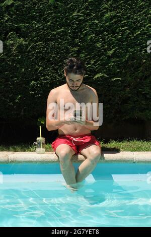 Young man using his smartphone sitting on the swimming pool edge. Summer concept. Stock Photo