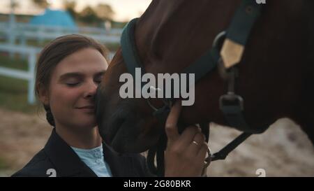 Horsewoman touching her face to the seal brown horse. Holding its bridle expressing her love for the horse. Sandy arena with wooden fence in the background. Bonding between horses and their owner. Stock Photo
