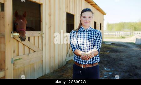 Horsewoman with horse feeds in her hands posing for the camera. Horse stable with stalls in the background. A dark brown horse looking out from the window of its stall. Horse stable daytime shot. Stock Photo