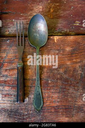 A tarnished antique fork with a wooden handle and a steel spoon lie on a worn, rustic wooden table. Stock Photo