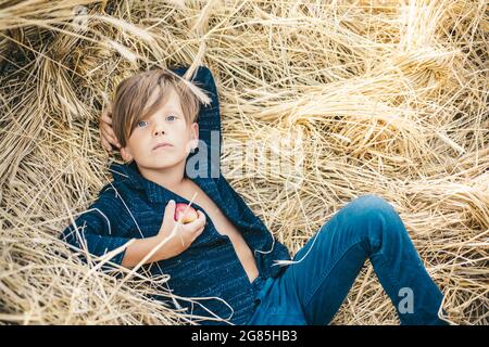 Fair-haired boy lies on hay background and eats an apple. Portrait of a cheerful boy lying in a hay. Little boy advertises natural products. Cute boy Stock Photo