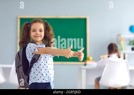 Adorable little girl with backpack showing thumbs up at classroom. Happy child going back to school, aiming for success Stock Photo