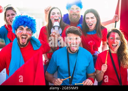 Football supporter fans friends cheering to soccer cup match at intenational stadium. Young people group with red and blue t-shirts having excited fun Stock Photo