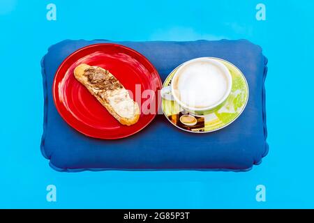 A cup of cappuccino and a red plate with a slice of bread spread with black and white chocolate cream on top, on an inflatable cushion that floats on Stock Photo