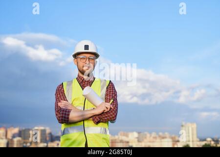 Portrait of caucasian bearded manager, architect or building contractor at sunny day with blue cloudy sky and urban city view. Man in white helmet and orange jacket standing on construction site Stock Photo