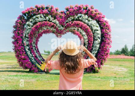 young woman in pink dress and hat stands on a background of flower arches. Image with a selective focus on hat. Heart-shaped wedding arch Stock Photo
