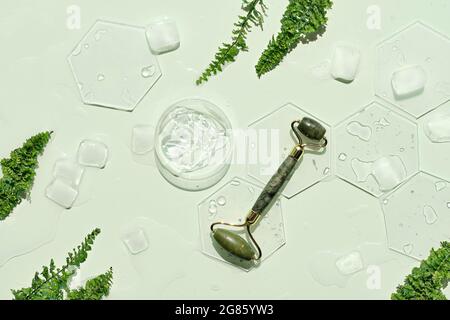 Self made moisturizer, green jade face roller with ice cubes. Exotic fern leaves on mint green background. Minimal flat lay, top view. Facial massage Stock Photo