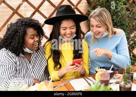 Young multiracial women watching on mobile smartphone while having healthy lunch in coffee brunch bar during corona virus outbreak Stock Photo