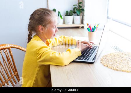 Caucasian blonde school girl, typing on a laptop keyboard, doing homework, blogging, chatting, viewing social media pages or playing. E-learning, back Stock Photo