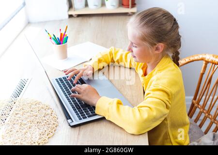 Caucasian blonde school girl, typing on a laptop keyboard, doing homework, blogging, chatting, viewing social media pages or playing. E-learning, back Stock Photo