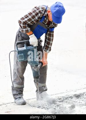 A worker repairs the road surface with a jackhammer on a summer day. Construction works on the road. Industrial background. Stock Photo