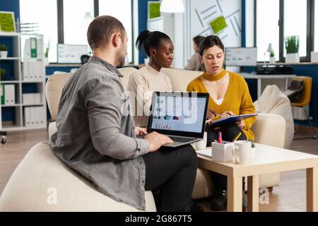 Businessman holding laptop with financial ghraphics while diverse employees talking explaining project plan reviewing paperwork sitting on couch. Multiethnic coworkers analysing financial reports Stock Photo