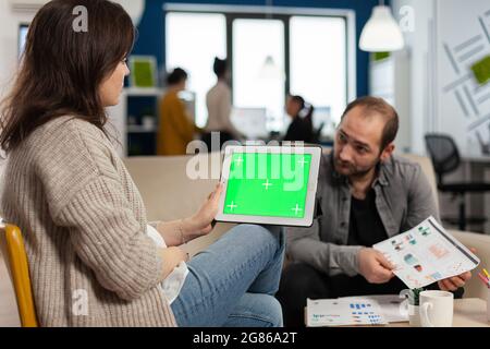Over the sholder of manager woman sitting on couch talking with coworker holding tablet with green screen while diverse team working. Multiethnic people planning project on chroma key display Stock Photo