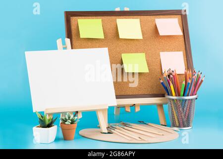Two wooden easel with blank canvas, cork board, brushes and colorful pencils on blue background. Stock Photo