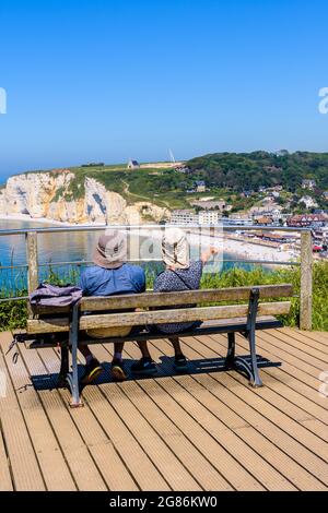 A couple of hikers sitting on a bench is enjoying the panoramic view over the beach town of Etretat and the Amont cliff in Normandy on a sunny day. Stock Photo