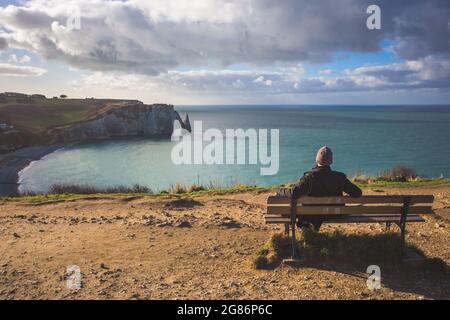 Tourist sitting on a bench admiring the seascape Etretat Aval cliff, rocks and natural arch landmark and blue ocean. Normandy, France Stock Photo