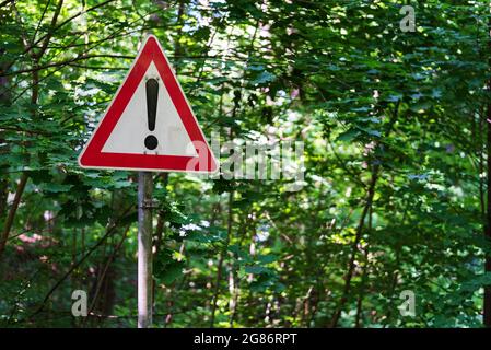 red and white warning sign with with exclamation mark against green trees Stock Photo