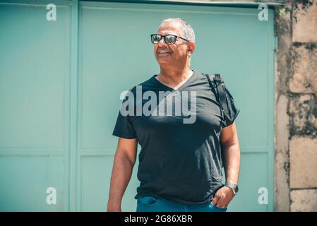 Smiling mature man standing in front of an old building holding his black backpack, looking away to side. Senior lifestyle travel. Stock Photo