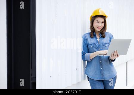 Beautiful Asian female engineer in jeans dress wearing yellow safety hard hat using laptop computer notebook doing job at construction site outside of Stock Photo