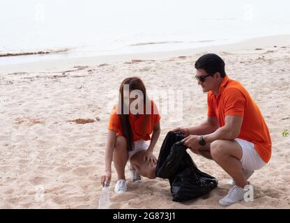 A young couple of volunteers cleans plastic and garbage in a black trash bag on a tropical beach. Stock Photo