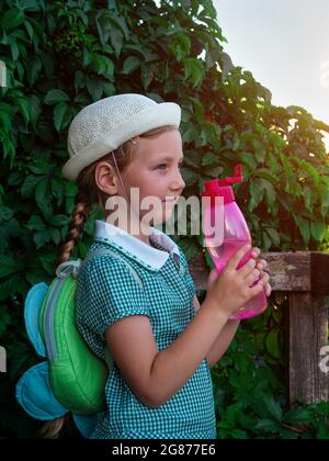 Cute school little girl drinks water from reusable pink bottle outdoor.  Child in hat enjoys fresh cold water on green summer street. Body  rehydration Stock Photo - Alamy