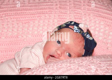 Close up portrait of a newborn baby girl with brown hair wearing floral headband and pink striped onsie tshirt. Laying on tummy on a pink crochet blan Stock Photo
