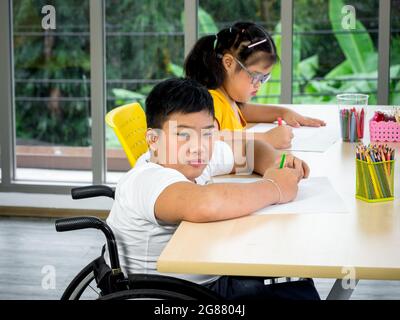 A disabled child in a wheelchair and a cute little girl with down syndrome is learning about drawing and painting while enjoying development exercises Stock Photo