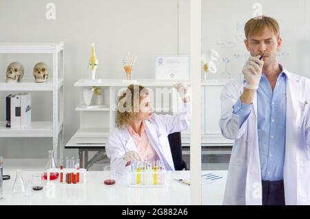 Young brazilian scientist write chemical formulas on the glass board. Young blonde assistant looking at the color of chemicals to observe the acidity Stock Photo