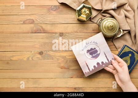 Female hand with greeting card for Eid al-Adha (Feast of the Sacrifice), Quran and lantern on wooden background Stock Photo