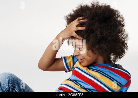 Smiling preschooler african boy wearing t-shirt sitting over white wall, side view Stock Photo