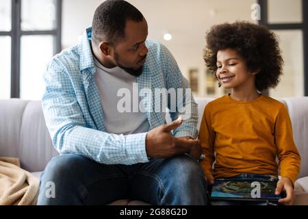 Black father and son talking and using tablet computer while sitting on sofa at home Stock Photo