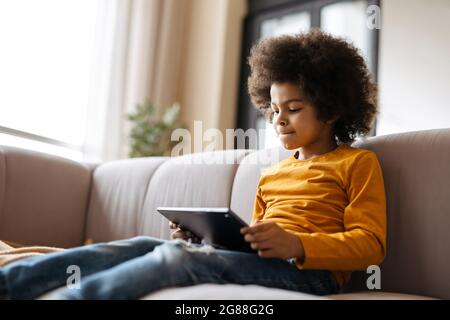Black curly boy using tablet computer while sitting on sofa at home Stock Photo