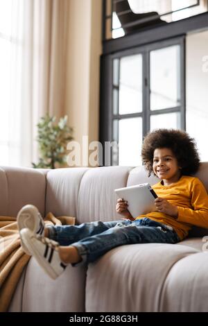 Black curly boy smiling and using tablet computer while sitting on sofa at home Stock Photo