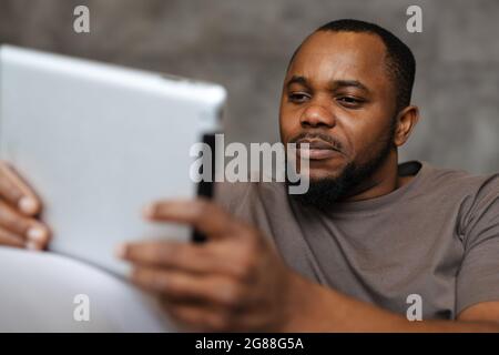 Black unshaven man using tablet computer while sitting on sofa at home Stock Photo