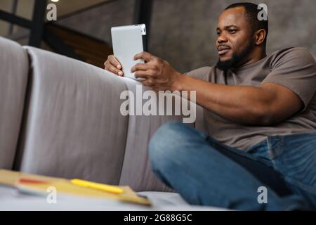 Black unshaven man using tablet computer while sitting on sofa at home Stock Photo