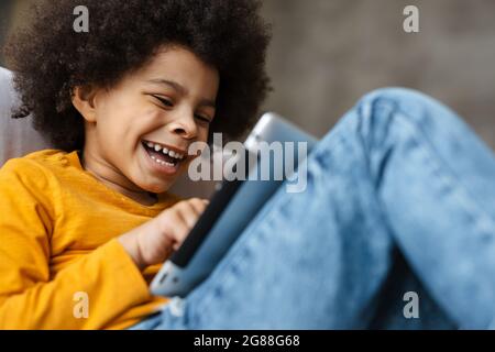 Black curly boy smiling and using tablet computer while sitting on sofa at home Stock Photo
