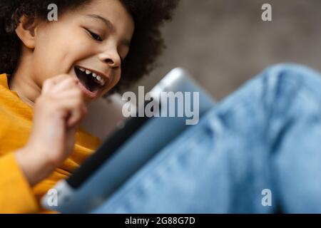 Black curly boy smiling and using tablet computer while sitting on sofa at home Stock Photo