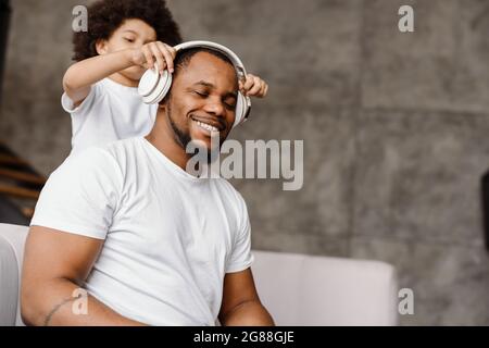 Black father and son smiling and using headphones while sitting on sofa at home Stock Photo