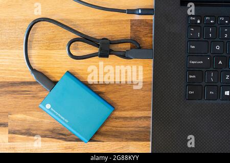 A blue Samsung 500GB encrypted solid state drive (SSD) external portable drive for data backup with a USB-C connection cable connecting it to a laptop Stock Photo