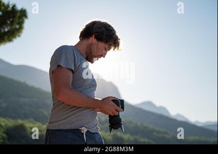 Young male photographer checking the settings on display of his dslr camera, standing outside on a beautiful summer morning. Stock Photo