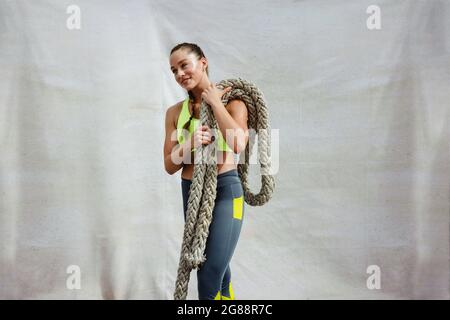 fitness woman in exercise outfit lifting battle rope and standing on white background. Sportswoman with battle rope relaxing after doing workout routi Stock Photo