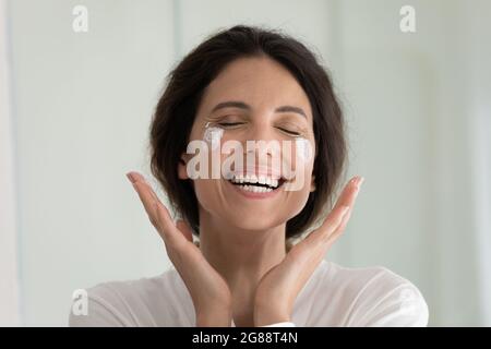 Overjoyed millennial latina woman feel pleasure applying cream on face Stock Photo