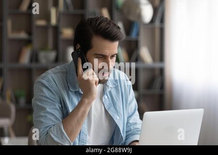 Concentrated young man talking on phone, working on computer. Stock Photo