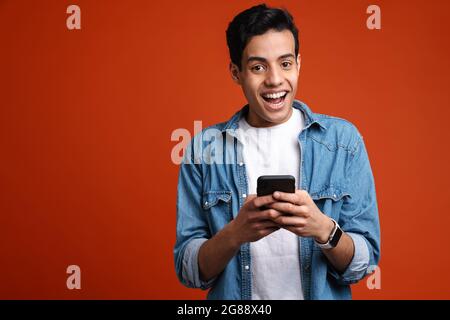 Brunette hispanic man in shirt smiling and using cellphone isolated over red background Stock Photo